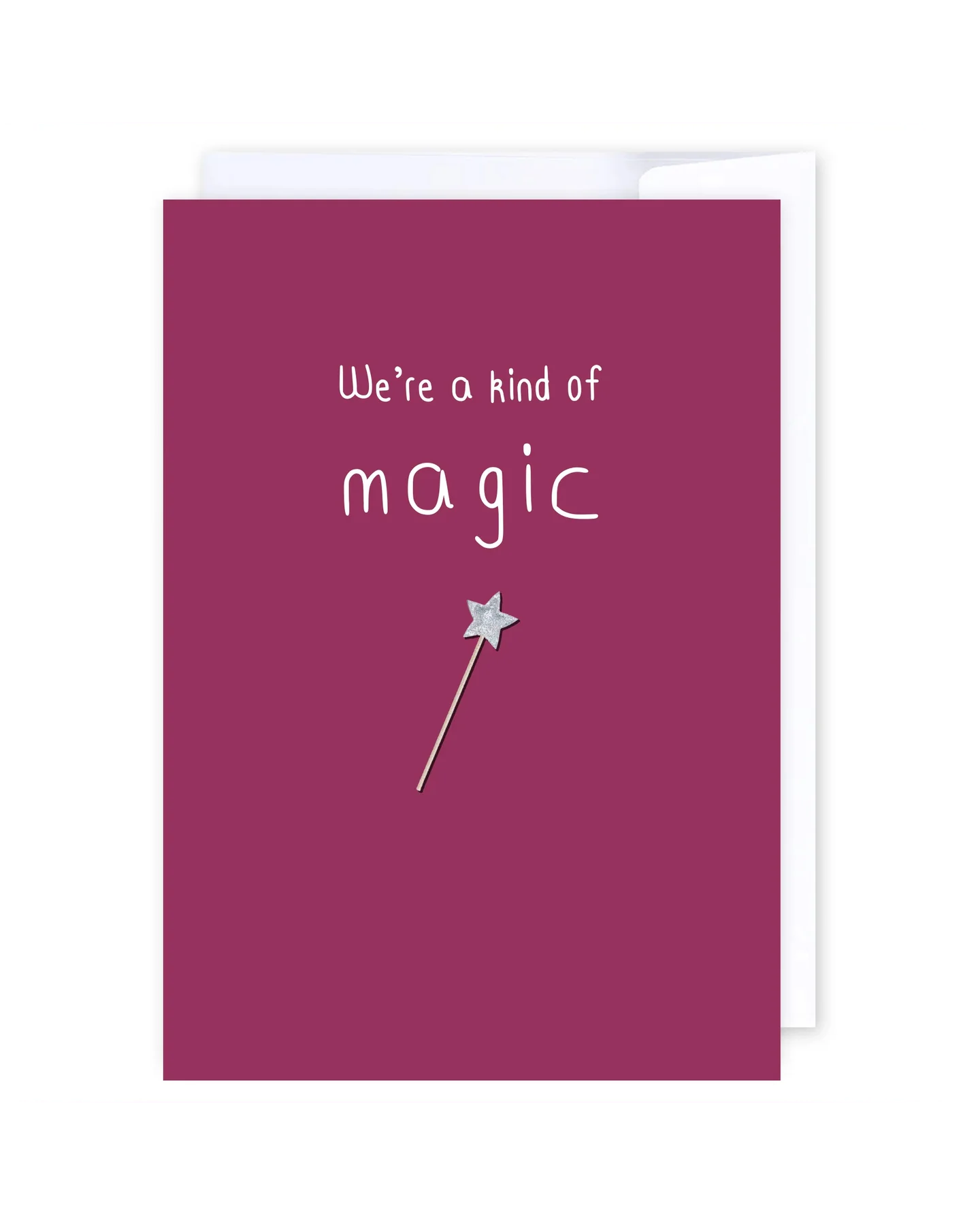 A maroon, New Zealand-made greeting card named “Were a kind of magic,” featuring white text that reads “We’re a kind of magic” and a silver star wand illustration beneath the text. A blank card is shown with part of its envelope visible behind it.