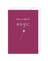 A maroon, New Zealand-made greeting card named “Were a kind of magic,” featuring white text that reads “We’re a kind of magic” and a silver star wand illustration beneath the text. A blank card is shown with part of its envelope visible behind it.